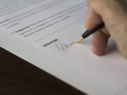Close-up photo of a hand holding a black ballpoint pen and signing a printed legal document